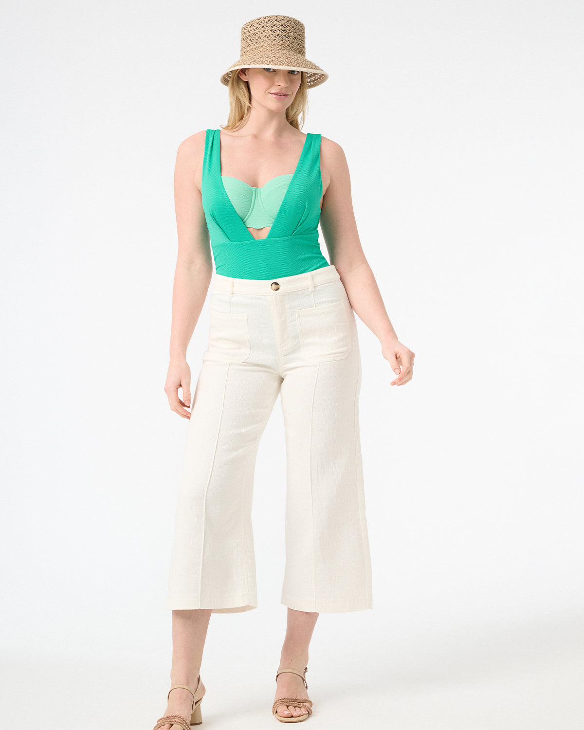 A model is wearing a green one-piece swimsuit with a deep v-neckline and sleeveless design and white long pants and a hat