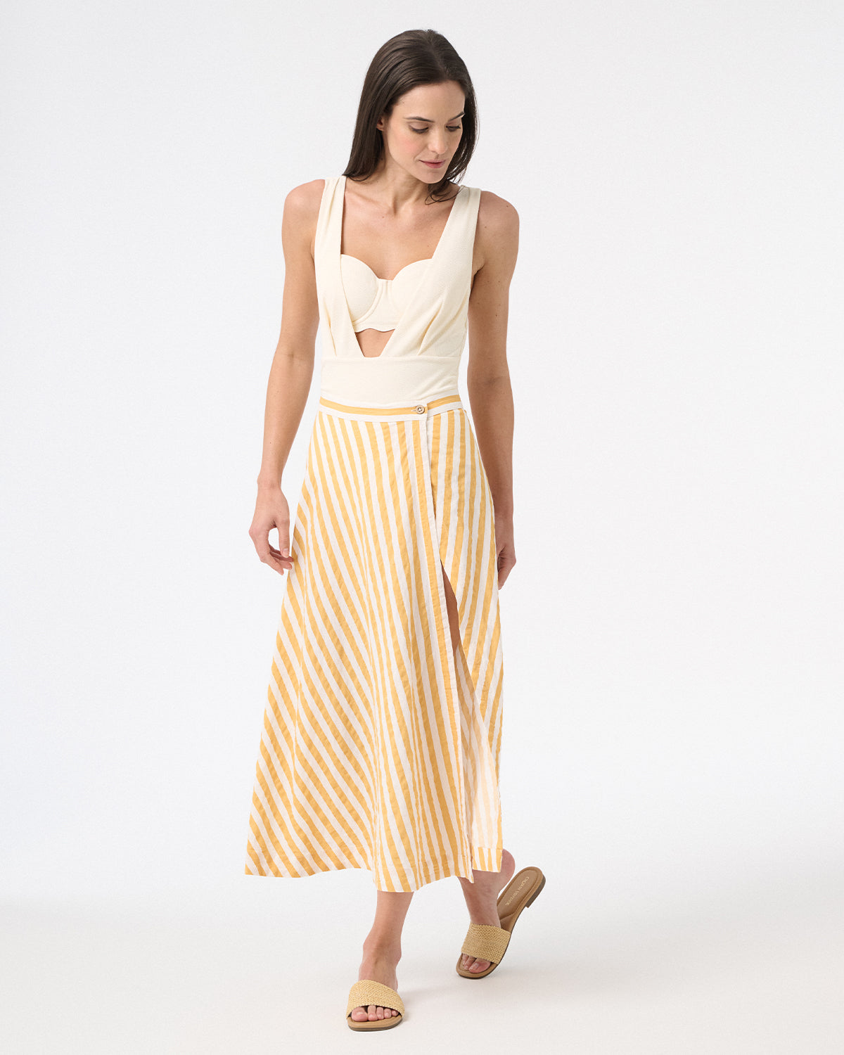 A model is wearing a cream one-piece swimsuit with a deep v-neckline and sleeveless design, withe a yellow and white long skirt over top and yellow sandals
