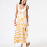 A model is wearing a cream one-piece swimsuit with a deep v-neckline and sleeveless design, withe a yellow and white long skirt over top and yellow sandals
