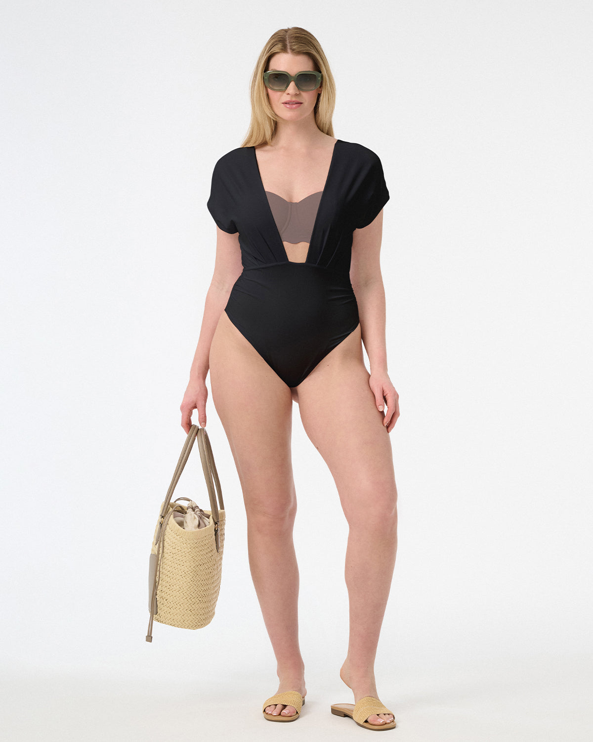 Woman wearing plunge neckline one piece black swimsuit with capped sleeves carrying bag, with yellow sandals and green sunglasses