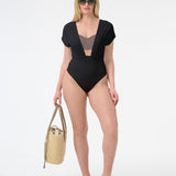 Woman wearing plunge neckline one piece black swimsuit with capped sleeves carrying bag, with yellow sandals and green sunglasses