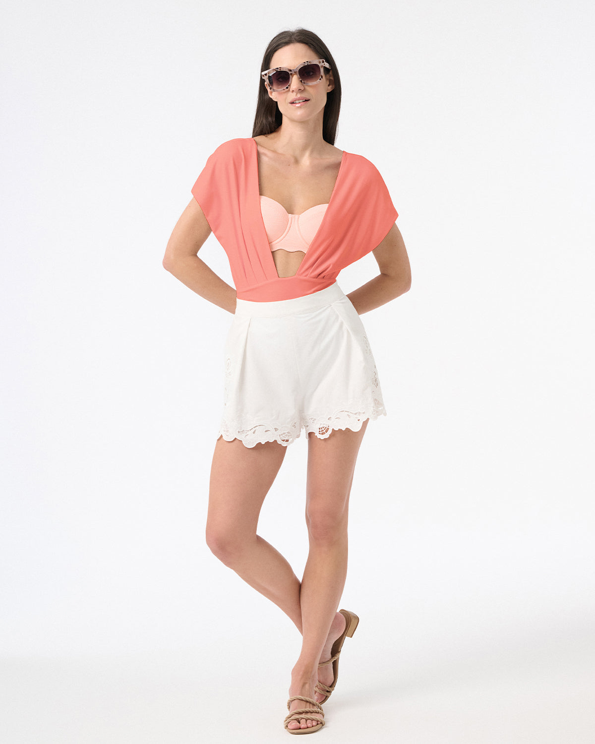Woman wearing one piece swimsuit with deep plunging neckline and white shorts with sandals and sunglasses