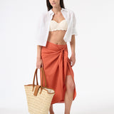 A woman wearing peach wrap skirt with cream bikini top, carrying a bag at her side with white button down shirt open in the front on top