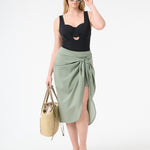 A woman wearing green wrap skirt with black swimsuit underneath carrying a bag at her side with green sunglasses on her face