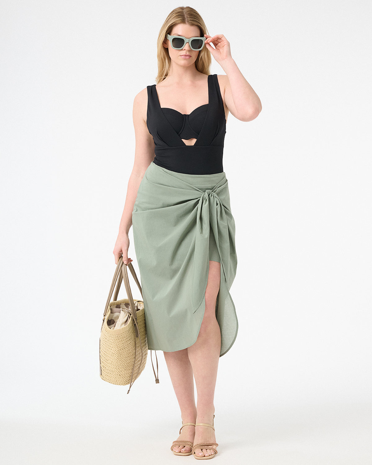 A woman wearing green wrap skirt with black swimsuit underneath carrying a bag at her side with green sunglasses on her face