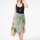 A woman wearing green wrap skirt with black swimsuit underneath carrying a bag at her side with green sunglasses on her face