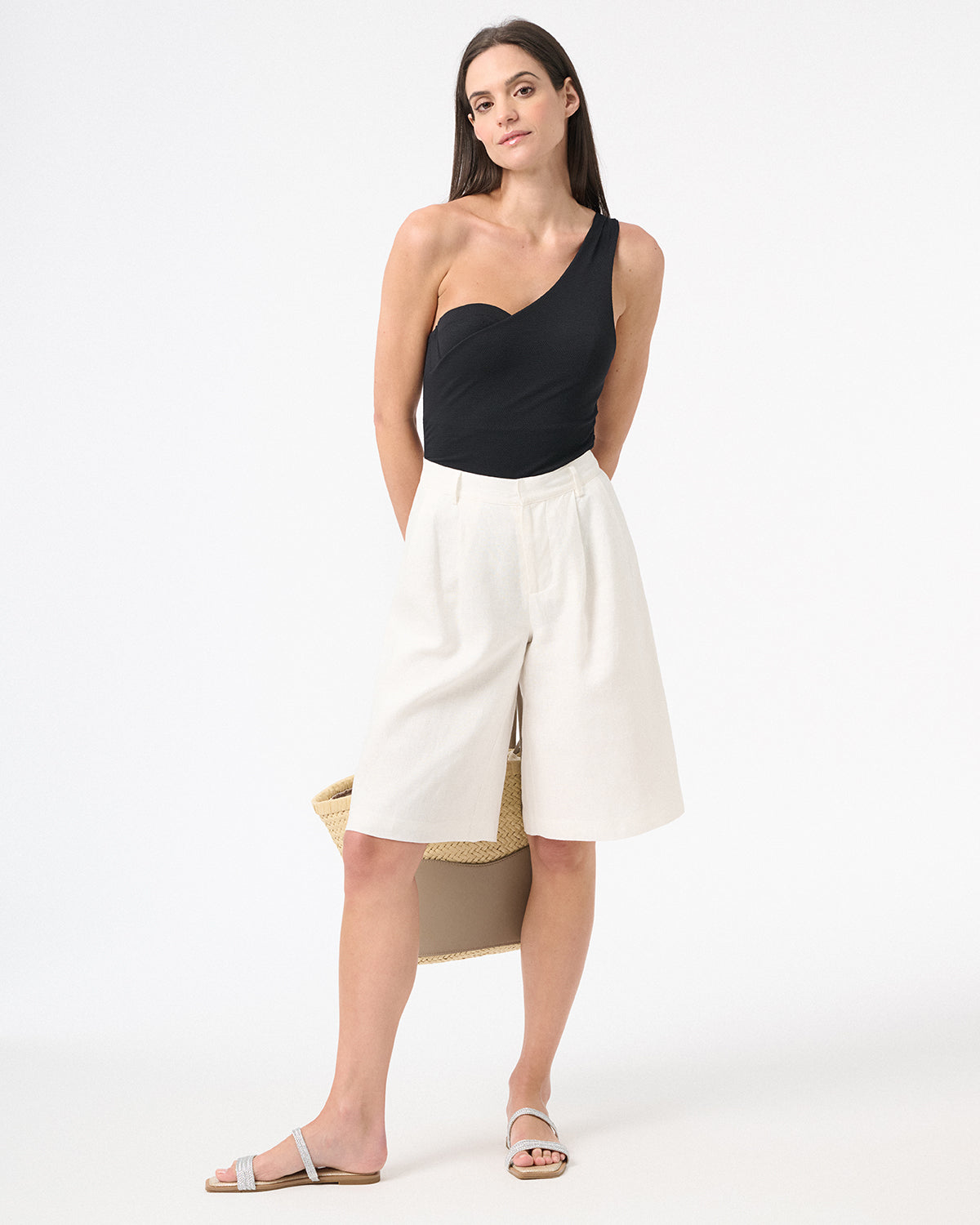 Woman wearing black one piece swimsuit with one shoulder design with long white shorts and carrying a bag behind her
