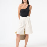Woman wearing black one piece swimsuit with one shoulder design with long white shorts and carrying a bag behind her